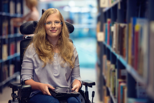 Young woman sitting in wheelchair holding digital tablet, looking at camera and smiling.