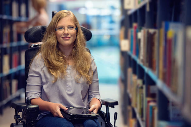 Portrait of young woman in wheelchair in library Young woman sitting in wheelchair holding digital tablet, looking at camera and smiling. physical disability photos stock pictures, royalty-free photos & images
