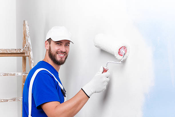 painter in white dungarees, blue t-shirt Portrait of handsome young smiling painter in white dungarees, blue t-shirt, cap and gloves painting a wall with paint roller, looking at camera house painter stock pictures, royalty-free photos & images