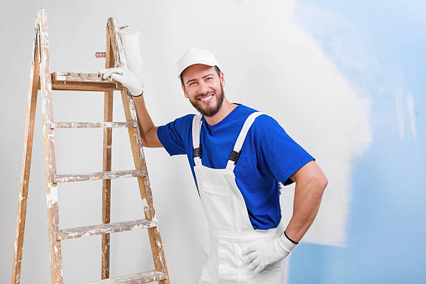 painter in white dungarees, blue t-shirt Portrait of handsome young smiling painter in white dungarees, blue t-shirt, cap and gloves leaning on a vintage ladder with paint roller, looking at camera house painter ladder paint men stock pictures, royalty-free photos & images