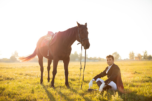 Man sitting next to his horse on meadow at sunset and looking away. Man looks very stylish with black tuxedo, white pants and black leather boots. 
