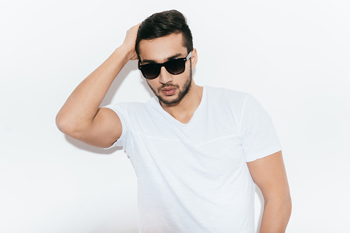 Handsome young Indian man in sunglasses holding hand in hair and looking at camera while standing against white background