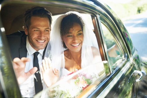 Shot of a newlywed couple looking out the window of a car and waving