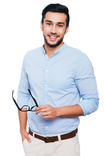 Confident young Indian man carrying his eyeglasses and smiling while standing against white background
