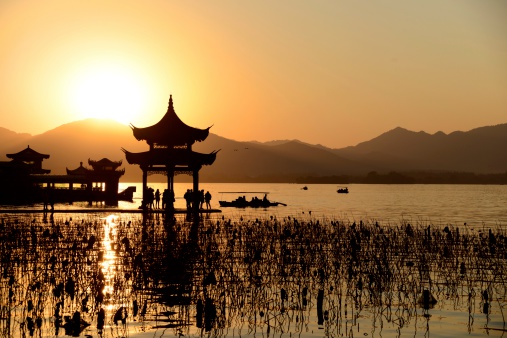Chinese temple in west lake at sunset, small ships in the water. The sun sinks behind the mountain. Clear Sky.