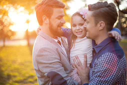 Young gay parents with their daughter having fun in park. Parents holding girl in arms. Enjoying in beautiful sunset. Caucasian ethnicity.