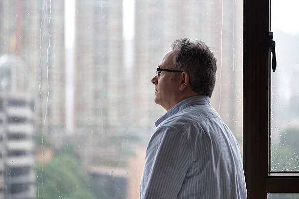 Serious Man Looking out Window on Rainy Day A mature man stares out the window at offices and apartment buildings in the background. Rain drops and streaks can be seen on the window.  Hong Kong. seasonal affective disorder stock pictures, royalty-free photos & images