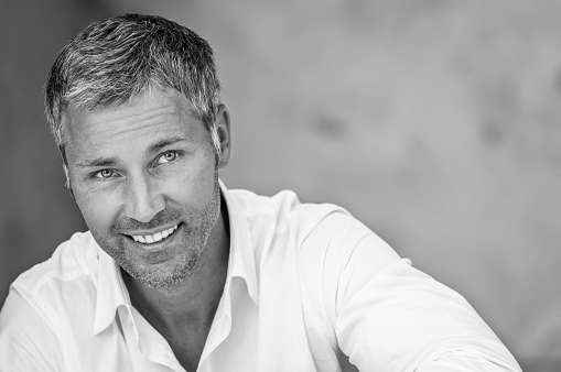 An attractive masculine man with grey hair looking friendly. He is sitting on the ground, leaning with his shoulder at a wall and smiling. He is wearing a white button down shirt with open collar. The black and white image was shot outdoors. There is some copy space on the top of the right side.