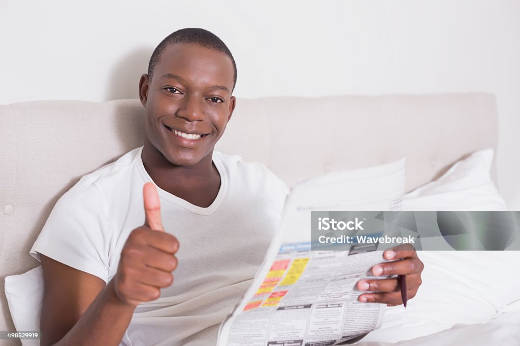 Happy man sitting up in bed with newspaper Happy man sitting up in bed with newspaper giving thumbs up at home in bedroom 20-29 Years Stock Photo