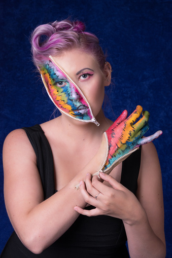 Vertical colour studio shot of young woman with pink hair in 40s style and zippers on face and hand. Zippers are open to show colourful abstract rainbow makeup. Front view, looking at camera while appearing to pull open zipper on hand with other hand. Waist up on dark blue crushed velvet background.