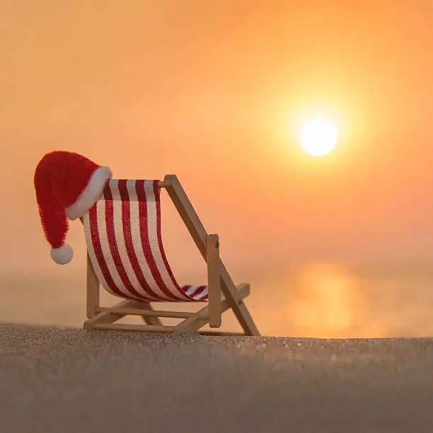 Striped red-white deckchair with christmas santa hat at ocean sandy beach during sunset, xmas or New Year's vacation in hot countries concept