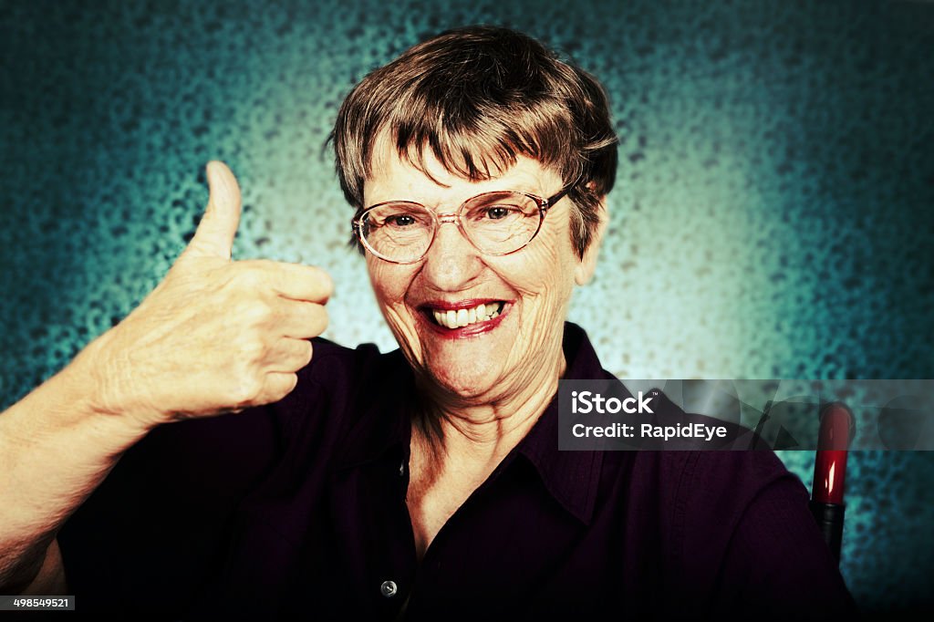 Happy old woman gives cheerful, approving thumbs-up signal A wrinkled old woman, seated in a wheelchair, smiles and gives a happy thumbs-up signal; her life is good! Patterned background. Senior Women Stock Photo