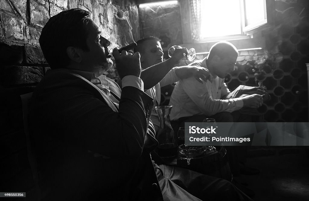 Three businessmen resting after a hard day at work. Three businessmen sitting in the wine cellar drinking wine and smoke cigar, resting after a hard day at work. Black and White. Mafia Stock Photo