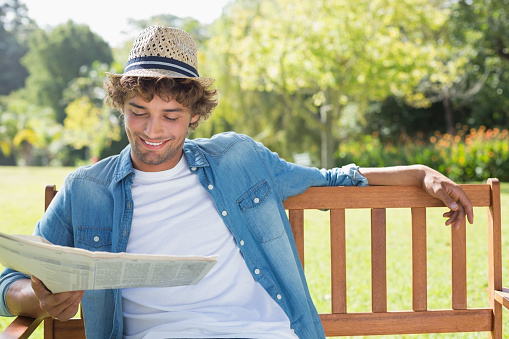 Hip young man sitting on park bench reading newspaper on a sunny day