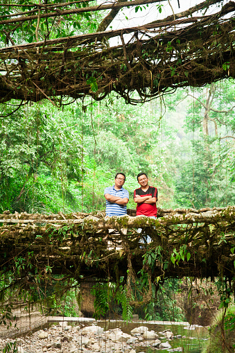 Two male tourists stand on double bridges made of old roots and vines hanging over a stream in northeastern India.  The old bridges are still traveled by villagers today to cross through the forest, woodland area. Nature backgrounds. 