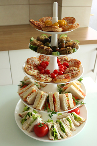 Photo showing a typically British afternoon tea, with sandwiches, cheese scones, mini quiches and various different coloured tomatoes - all displayed on a tall, white tiered cake stand, ready to be taken into the garden and followed by a slice of carrot cake.