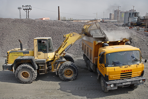 St. Petersburg, Russia - April 27, 2009: Construction equipment and machinery, a pile of construction gravel, yellow digger loads crushed stone into orange dumper truck tipper.