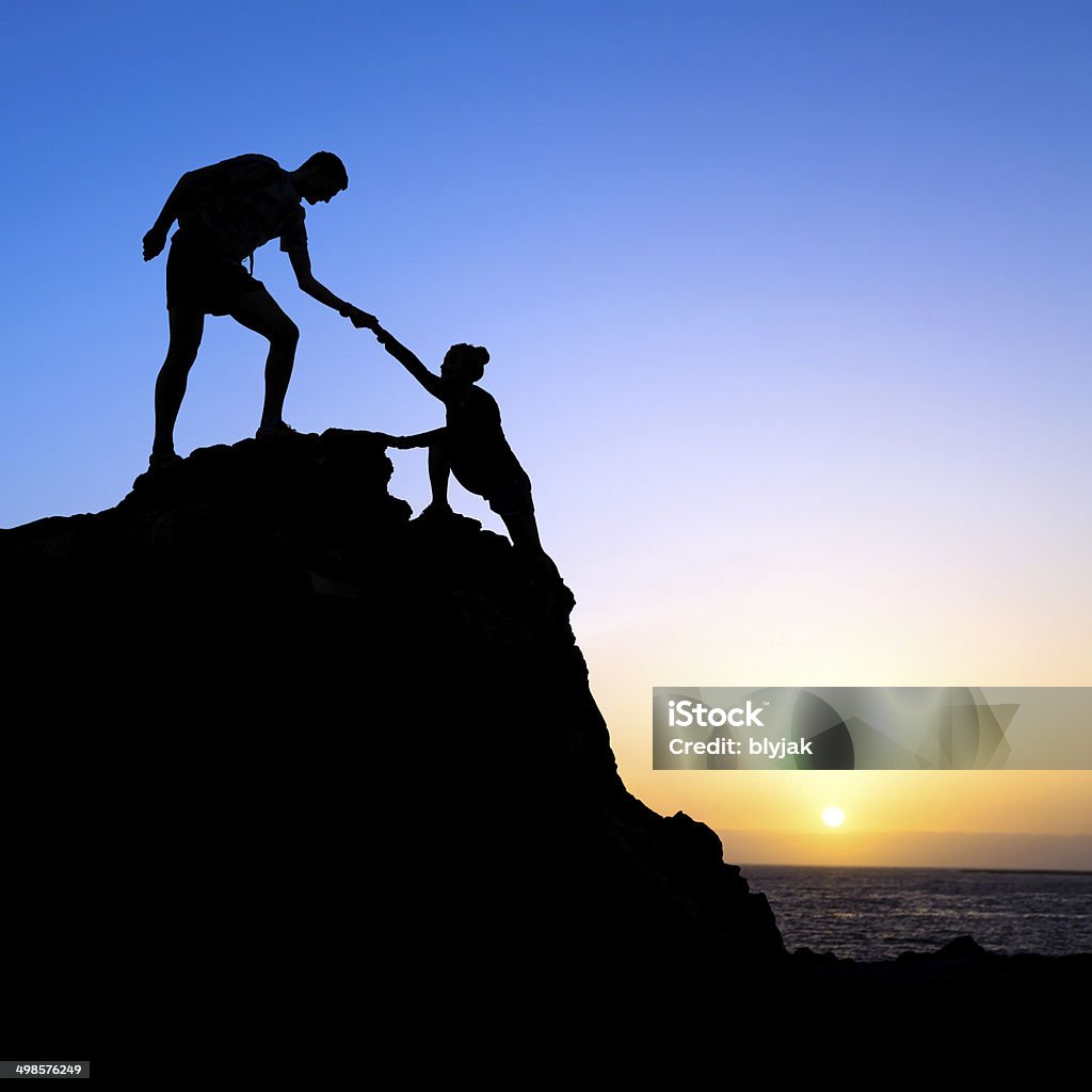 Man and woman help silhouette in mountains Couple hiking help each other silhouette in mountains, sunset and ocean. Male and woman hiker helping each other on top of mountain climbing, beautiful sunset landscape. A Helping Hand Stock Photo