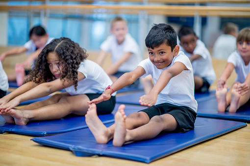 A multi-ethnic group of elementary age children are sitting on an exercise mat and are stretching forward to reach their toes.