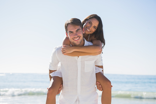 Handsome man giving girlfriend a piggy back smiling at camera at the beach