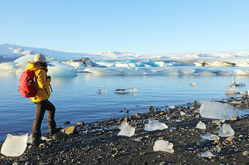 Female traveller is standing at the edge of Jokulsarlon glacial lagoon in Iceland on a beautiful sunny winter day. The hiker is wearing a warm yellow jacket and carrying a red backpack as she is looking at the drifting ice formations in the lake at the foot of a melting glacier which can be seen in the background,