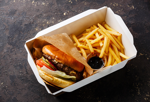 Burger with meat and French fries in paper box on dark background