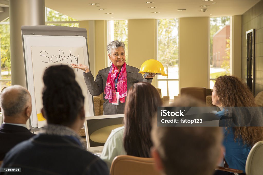 Safety in the workplace. Presentation with workers. African descent business woman gives safety presentation at office. Multi-ethnic group of professionals.  She holds construction hard hat. Occupational Safety And Health Stock Photo