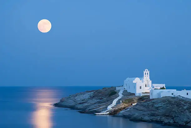A summer picture of the island Sifnos with the full moon and its reflections on the Aegean Sea. The famous church of Panagia Chrysopigi on the foreground.