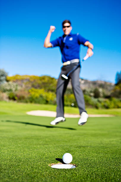 A young man playing jumps excitedly, gesturing in tirumph as his ball heads into the cup. It's a good golfing day!