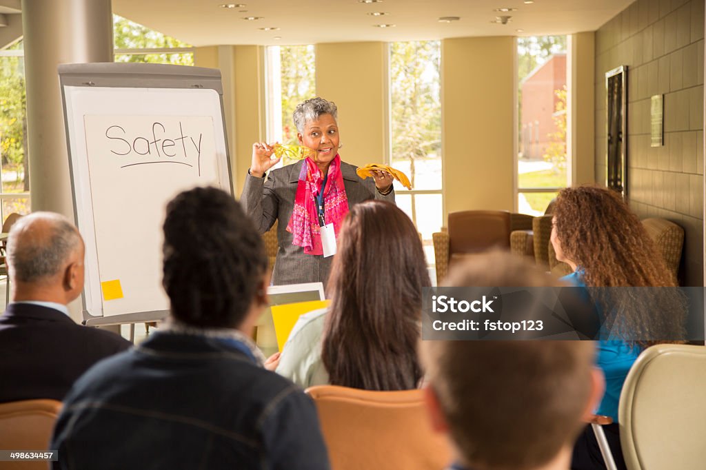 Safety in the workplace. Presentation with workers. African descent business woman gives safety presentation at office. Multi-ethnic group of professionals.  She holds safety glasses and gloves. Education Training Class Stock Photo
