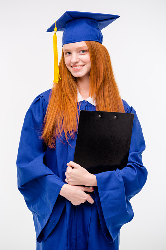 Portrait of pretty redhead cheerful graduated positive girl in blue gown and graduation cap holding clipboard