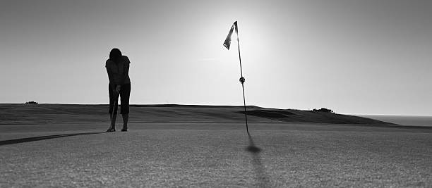 A woman golfer putting on the 18th hole - mono image with sun and sea in distance