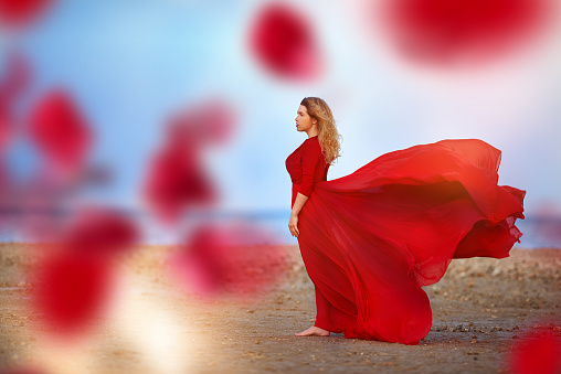 side view of blond hair woman on the beach, looking at view.flying red dress, flying red petals.