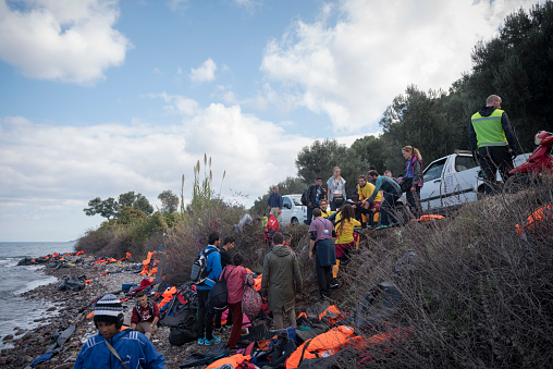 Lesbos, Greece - October 25, 2015: On the north coast of the Greek island of Lesbos, refugees and other migrants arriving by inflatable boat are helped up from the beach to the road, which they will follow to a registration center. Volunteers, including lifeguards, and a photographer are among the people standing on the road. So far in 2015, more than 500,000 migrants have crossed from Turkey to the Greek islands by boat.