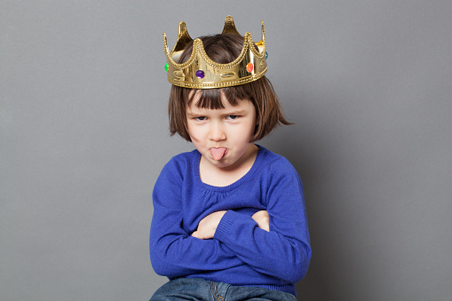 spoiled kid concept - cheeky preschool child with golden crown on head folding arms and sticking out tongue for disrespectful mollycoddled little king or queen metaphor,studio shot