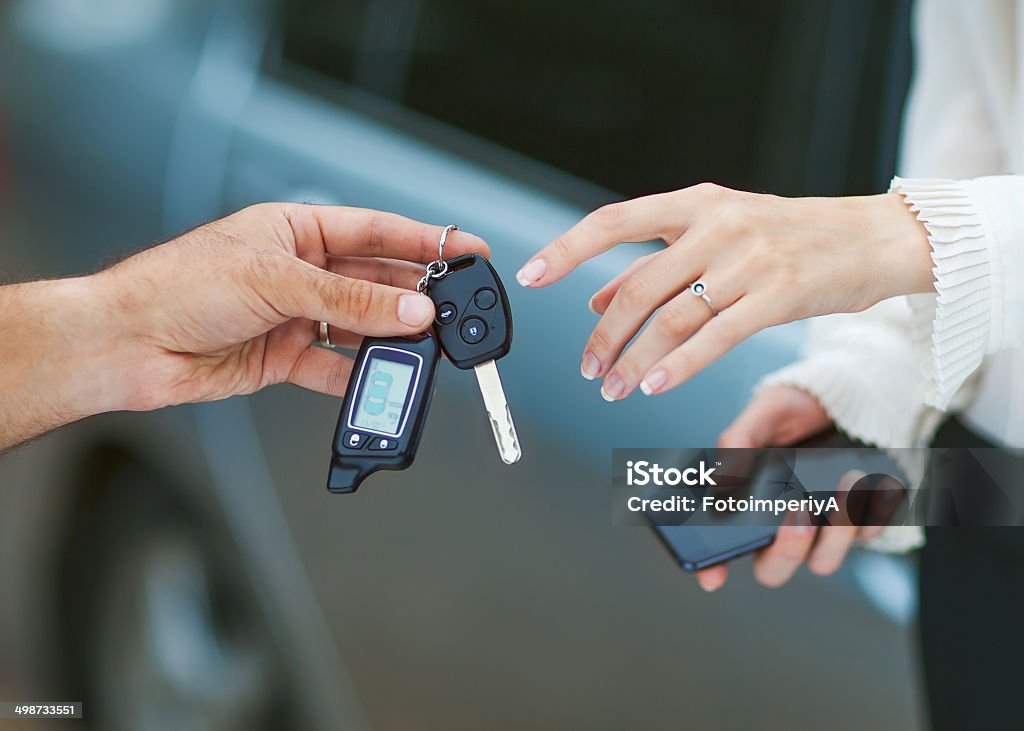 Male hand giving car key to female hand. Male hand giving car key to female hand. She is holding a cell phone. In the background, a fragment of the car. Adult Stock Photo