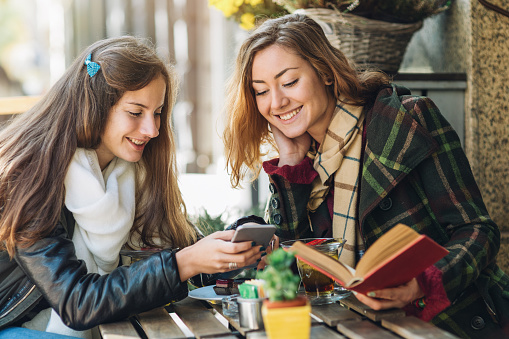 Two student girls drinking tea and enjoying warm autumn weather in a cafe. The girl on the left is showing something on her cell phone, while the girl on the right i holding old book. Casual autumn clothing.