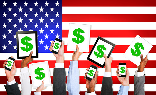 Multiethnic Group Of Business People Holding Technology Devices With A Dollar Sign On A Screen And American Flag As A Background