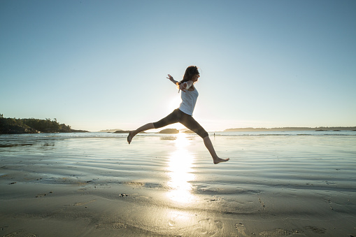 Cheerful young woman on the beach jumping high up for joy and freedom. The sunset is making a magic light reflecting on the sand.