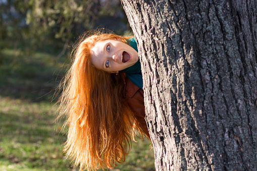 Amusing funny girl with beautiful long red hair looking out from the tree in park