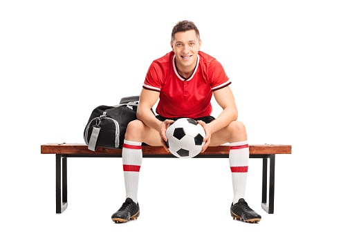Young football player holding a ball and sitting on a wooden bench isolated on white background