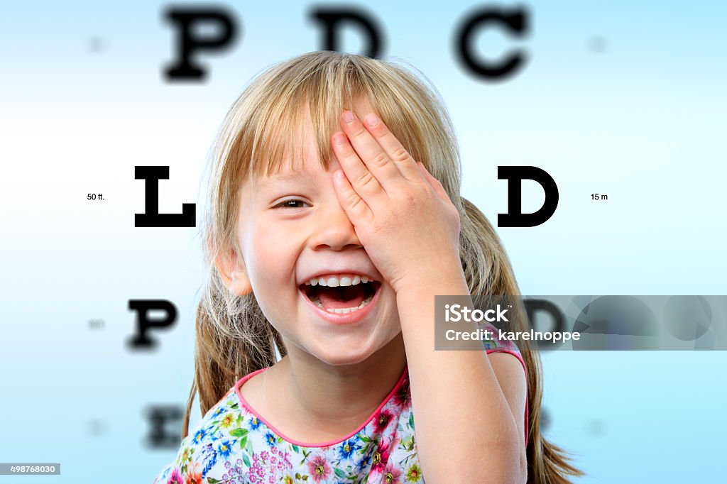 Girl having fun at vision test. Close up face portrait of happy girl having fun at vision test.Conceptual image with girl closing one eye with hand and block letter eye chart in background. Child Stock Photo