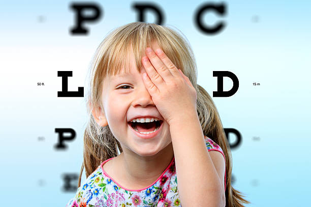 Girl having fun at vision test. Close up face portrait of happy girl having fun at vision test.Conceptual image with girl closing one eye with hand and block letter eye chart in background. myopia stock pictures, royalty-free photos & images