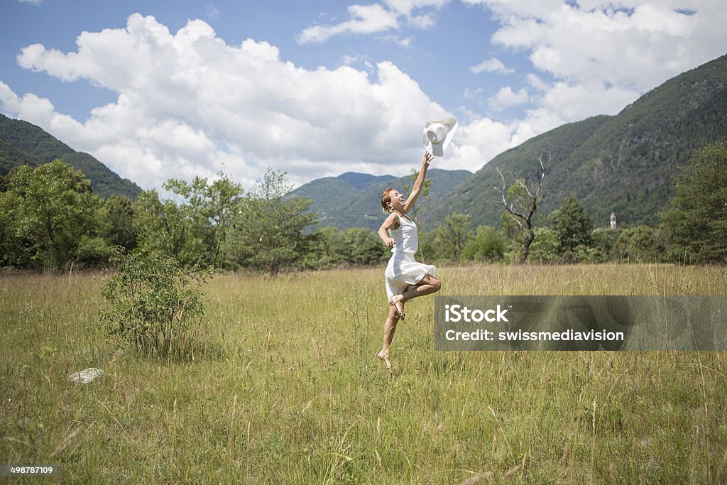 Young woman in field jumping and throwing hat in hair Young happy woman jumping and throwing her hat in the hair. Mountain and field landscape in Switzerland during a beautiful spring day. Active Lifestyle Stock Photo