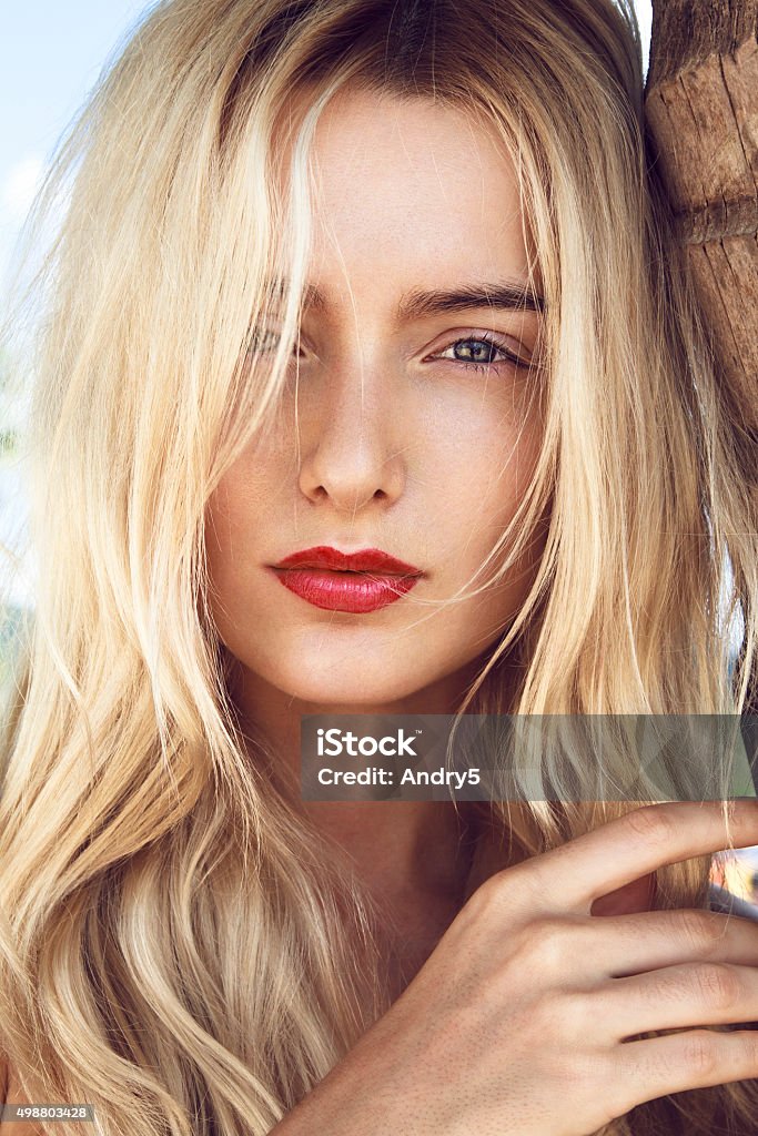 Around the palm Close-up portrait of beautiful young woman with curly long blonde hair, green mysterious eyes and red sexy big lips posing next to the palm and looking to you through lens. Women Stock Photo
