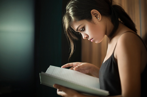Indoor low key image of a beautiful confident teenage Indian girl studying notes through her note book for the preparation of upcoming exams. She is at home in her bedroom wearing black spaghetti top with her back resting to the wooden door of her closet. Horizontal composition with copy space and selective focus.
