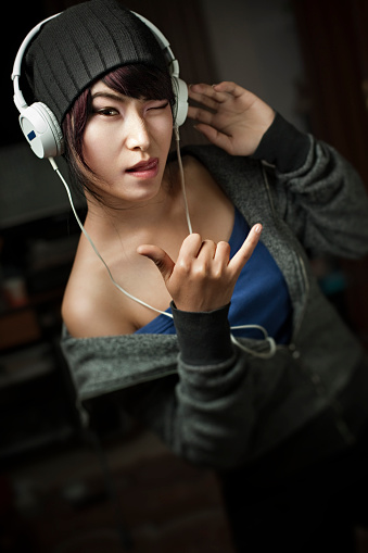 Indoor low key image of a fashionable Asian teenage girl listening music through headphones and dancing at night in her domestic room while giving a winking and tongue out gesture at camera. She is in off shoulder gray zipper jacket and blue top. Vertical, tilt composition with copy space and selective focus.