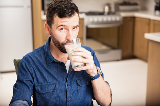 Portrait of a young handsome man with a beard drinking some milk from a glass and making eye contact
