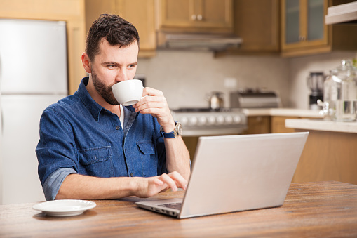 Portrait of a young busy man with a beard drinking some coffee and using a laptop computer to work from home