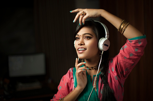 Indoor low key image of a fashionable Asian Hindu teenage girl listening music through headphones and dancing at night in her domestic room. She is in traditional Hindu dress that is Salwar Kameez and dupatta. Horizontal composition with copy space and selective focus.